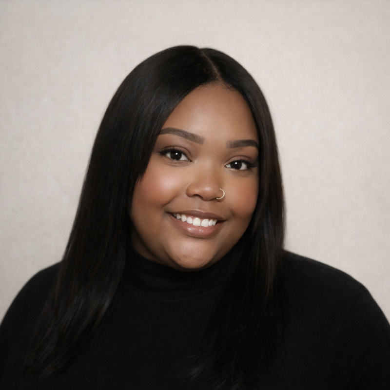 A young woman with dark hair, wearing a black top and a nose ring, smiling at the camera against a plain, light-colored background.