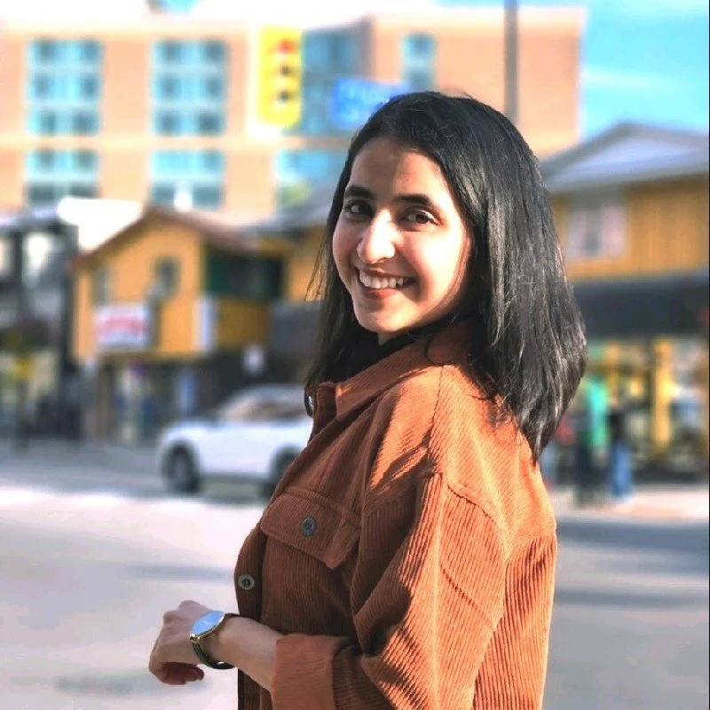 A smiling woman with shoulder-length black hair wearing a brown corduroy jacket standing outdoors in an urban setting with blurred buildings and cars in the background.