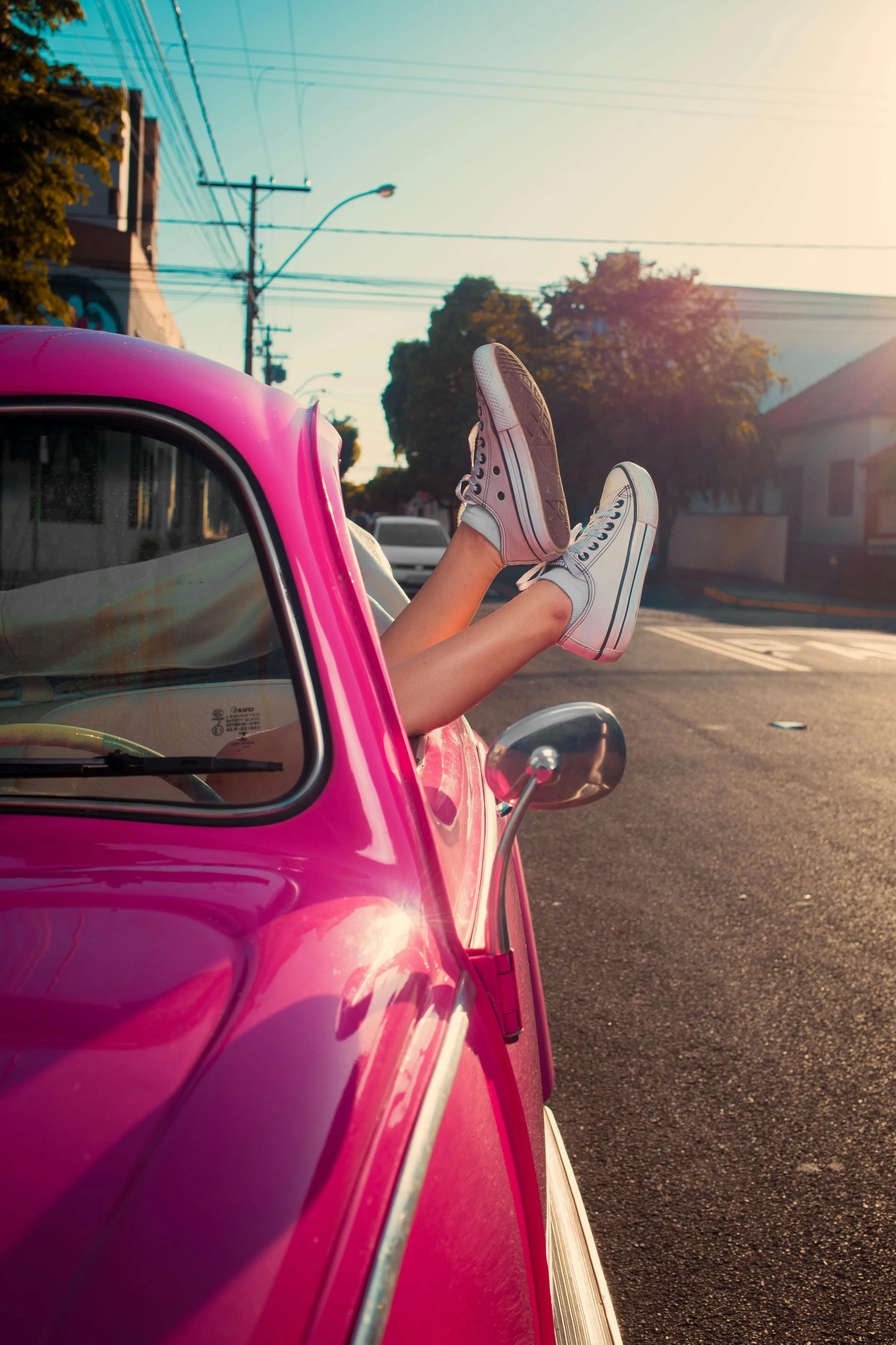 Person with legs extended out of a pink vintage car wearing white sneakers, parked on a city street during sunset.