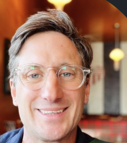 Close-up of a man with glasses smiling in a warmly lit indoor setting.