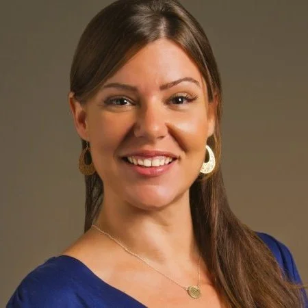 Portrait of a smiling woman with brown hair, wearing gold earrings, a blue top, and a gold necklace.