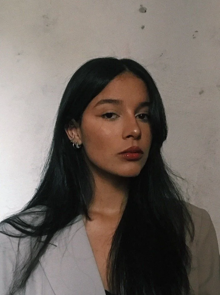 A young woman with long black hair, light makeup, red lipstick, and hoop earrings, looking at the camera against a plain wall.