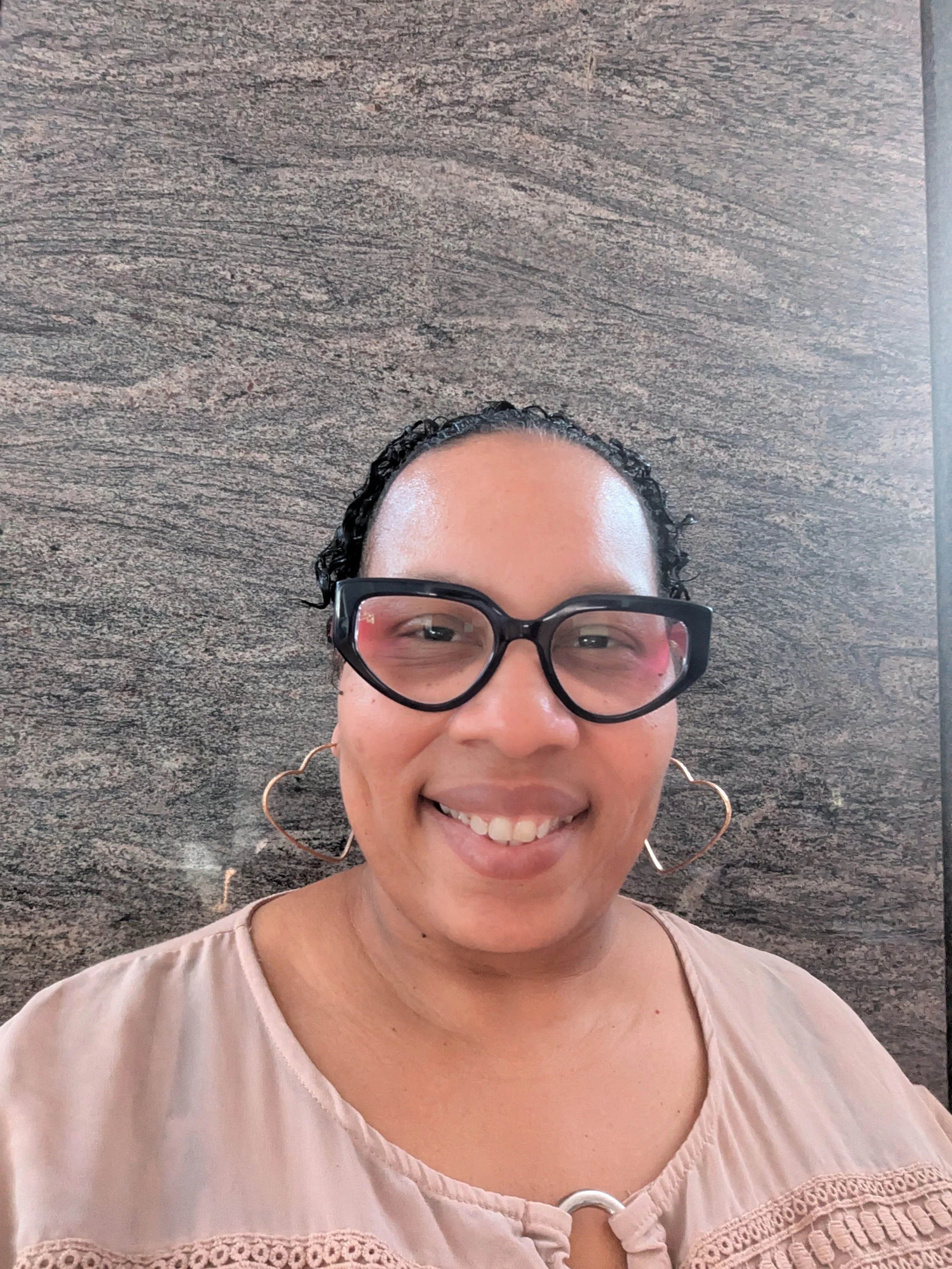 Smiling woman wearing black glasses and heart-shaped earrings, standing against a textured brown wall.