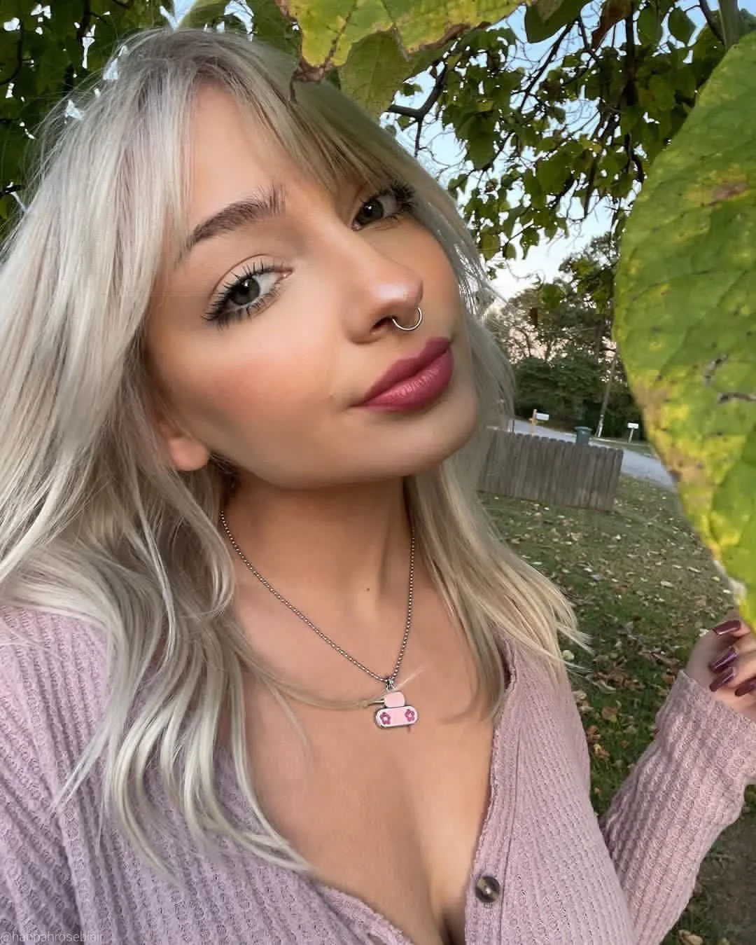 A young woman with platinum blonde hair, wearing a pink sweater and a necklace with a pink tank pendant, standing outdoors near green leaves, holding a branch with green-yellowing leaves.