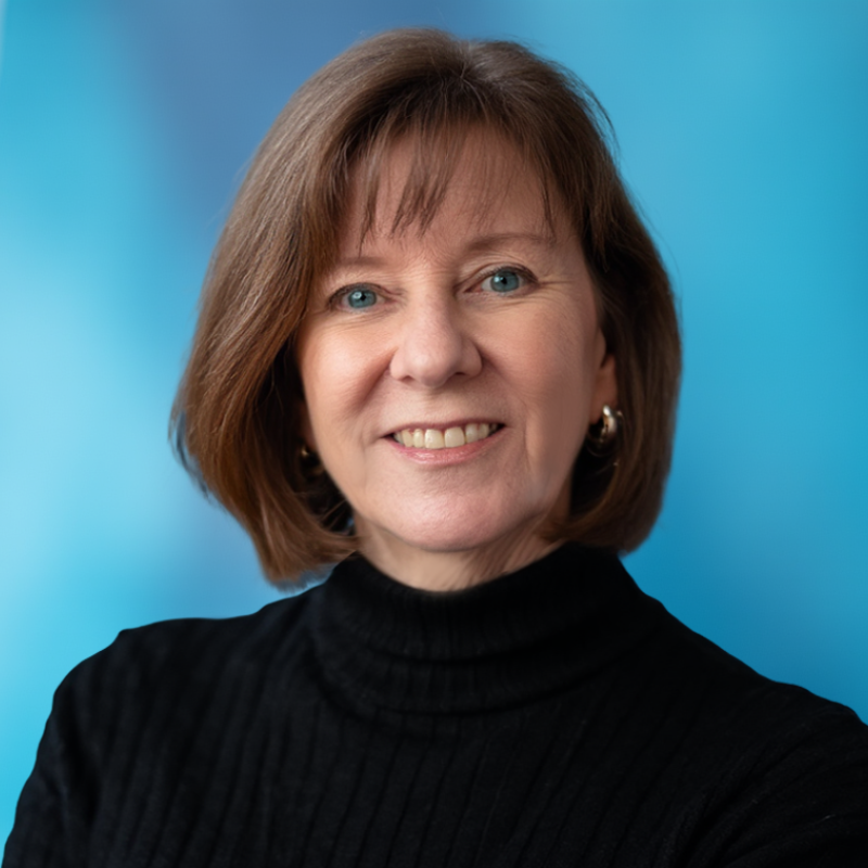 A middle-aged woman with shoulder-length brown hair and blue eyes, wearing a black turtleneck, smiling against a blue background.