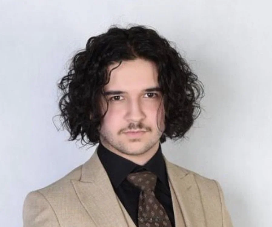 A man with curly dark hair, light skin, and a mustache, wearing a tan blazer, black shirt, and patterned tie, standing against a plain white background.