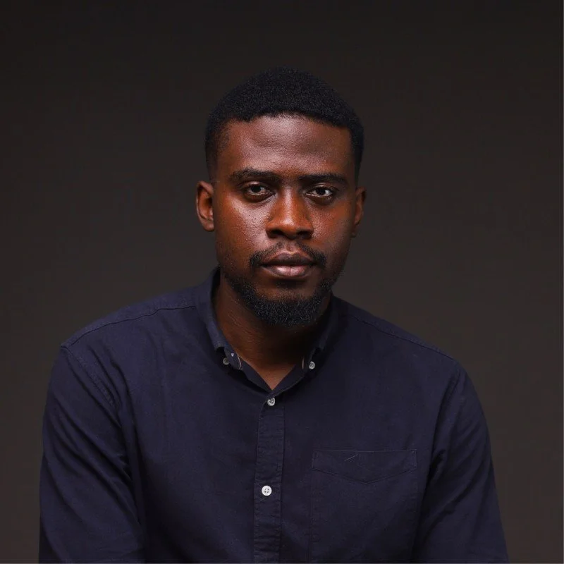 A portrait of a young African American man with short hair and a beard, wearing a dark button-up shirt against a plain dark background.