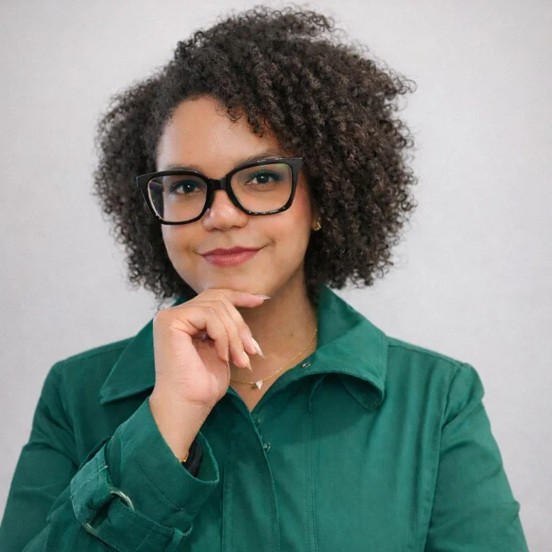 A woman with curly hair, wearing glasses and a green jacket, smiling with her hand resting under her chin.
