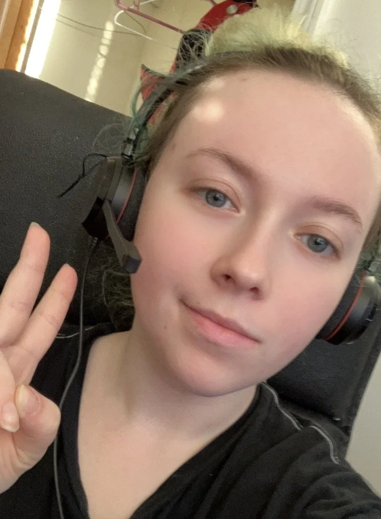 A young person with blue eyes and light skin taking a selfie while wearing a gaming headset and making a peace sign gesture with their right hand.