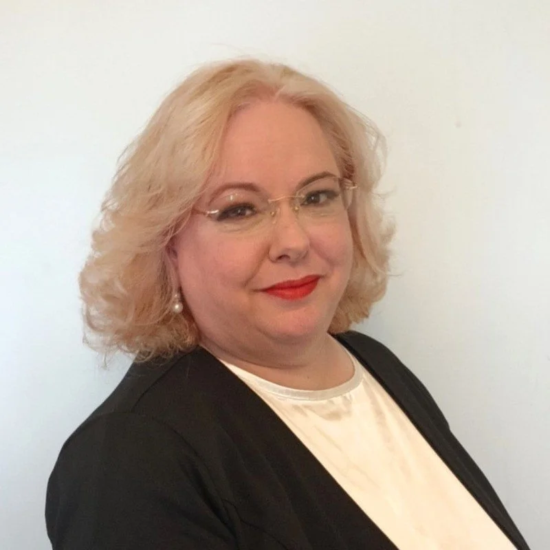 A woman with blonde, curly hair wearing glasses, pearl earrings, a white top, and a black blazer, smiling against a plain white background.
