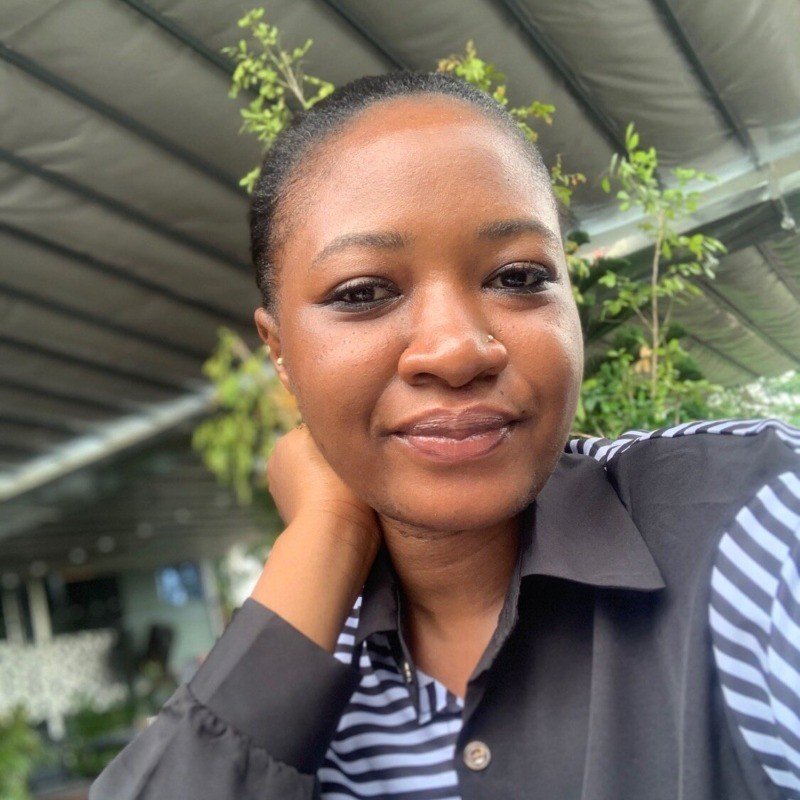 A young woman with dark skin and natural hair, smiling softly, resting her head on her hand, outdoors with greenery and a metal roof in the background.