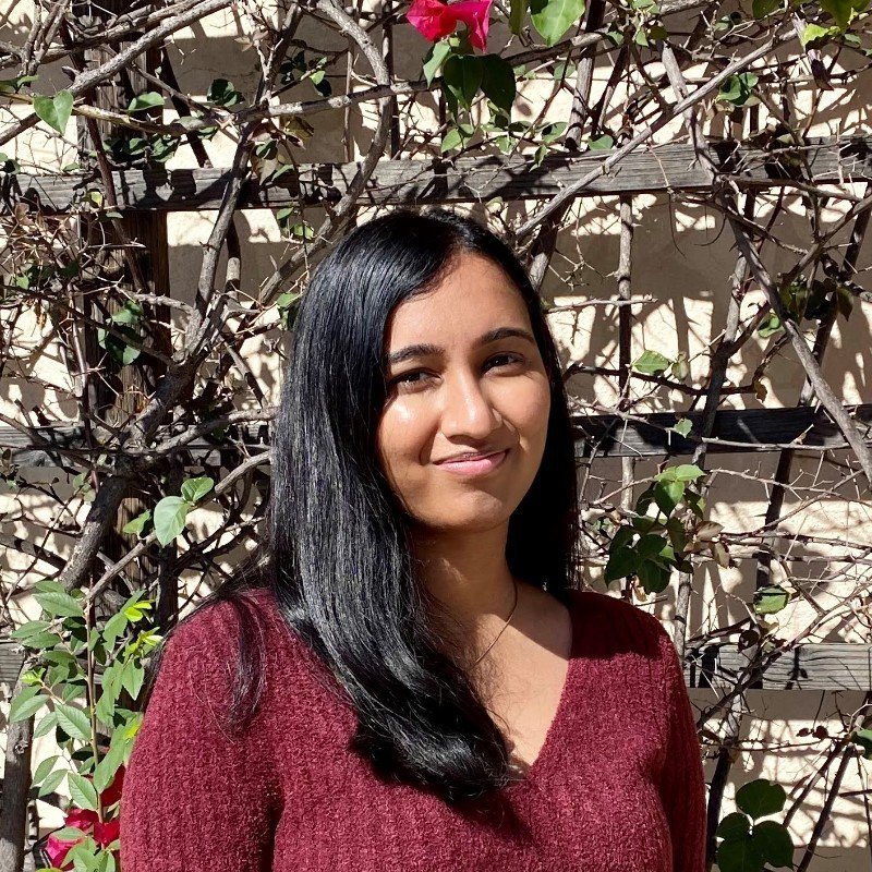 A woman with long black hair wearing a red sweater standing outside in front of a vine-covered wooden trellis with pink flowers.