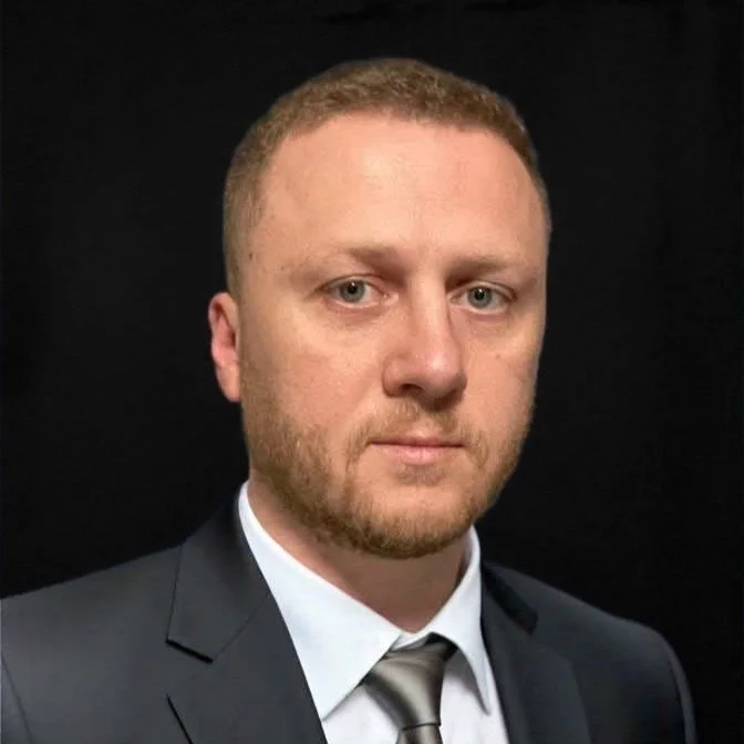 A man with short red hair and a beard dressed in a dark suit, white shirt, and silver tie against a black background.