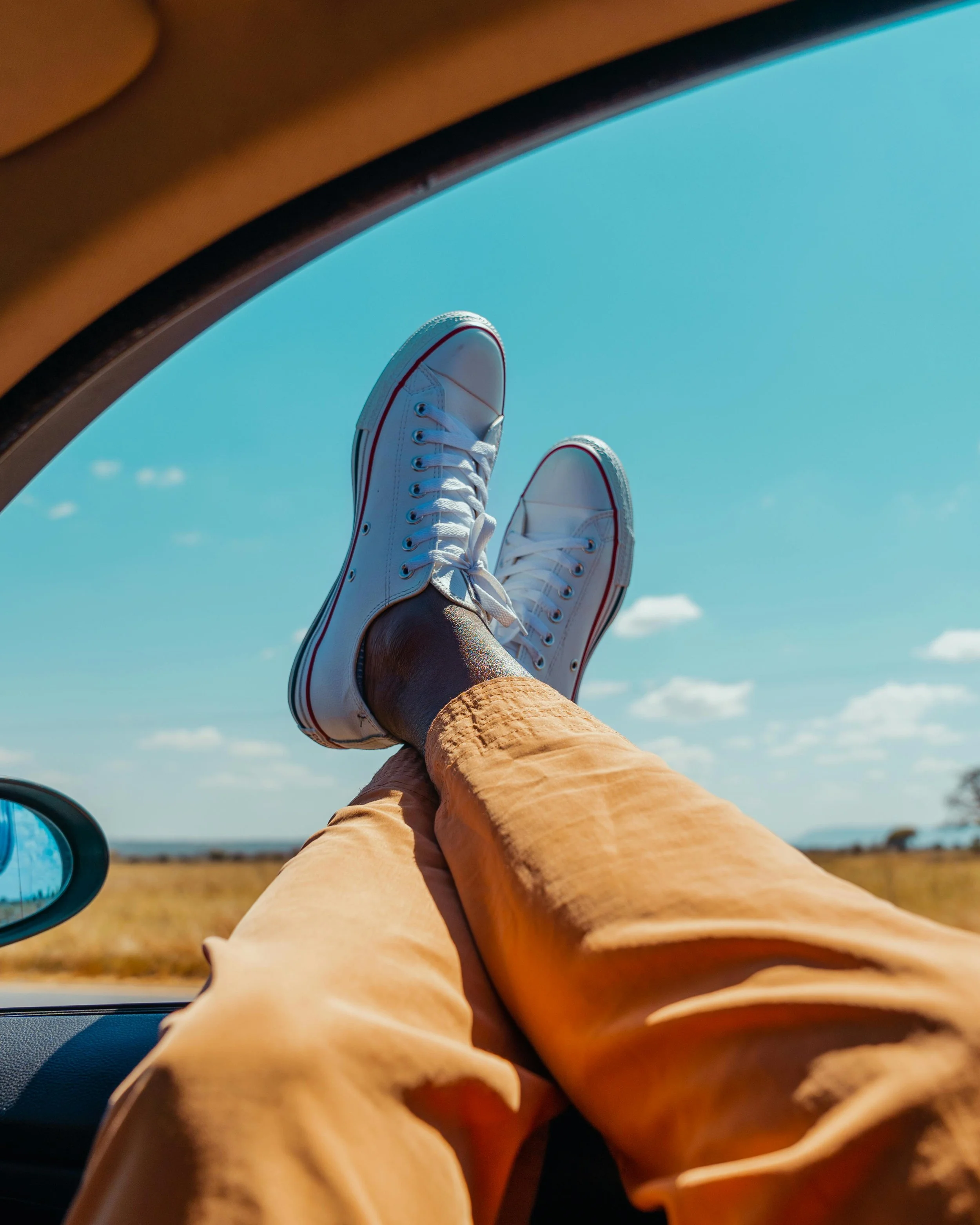 Person in yellow pants and white sneakers with red and black accents, relaxing with legs crossed out of a car window on a sunny day, blue sky and open fields in the background.