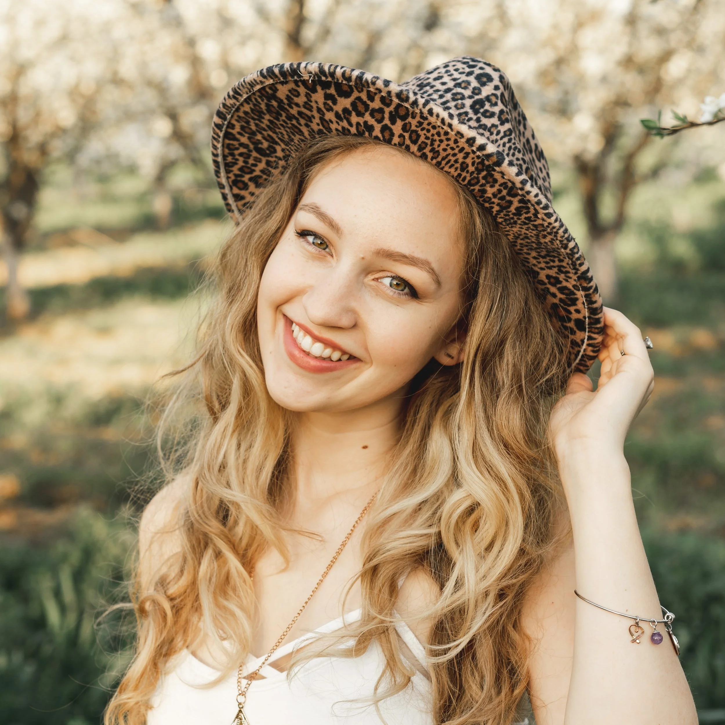 A young woman with long, wavy blonde hair wearing a leopard print hat, smiling outdoors with trees in the background.