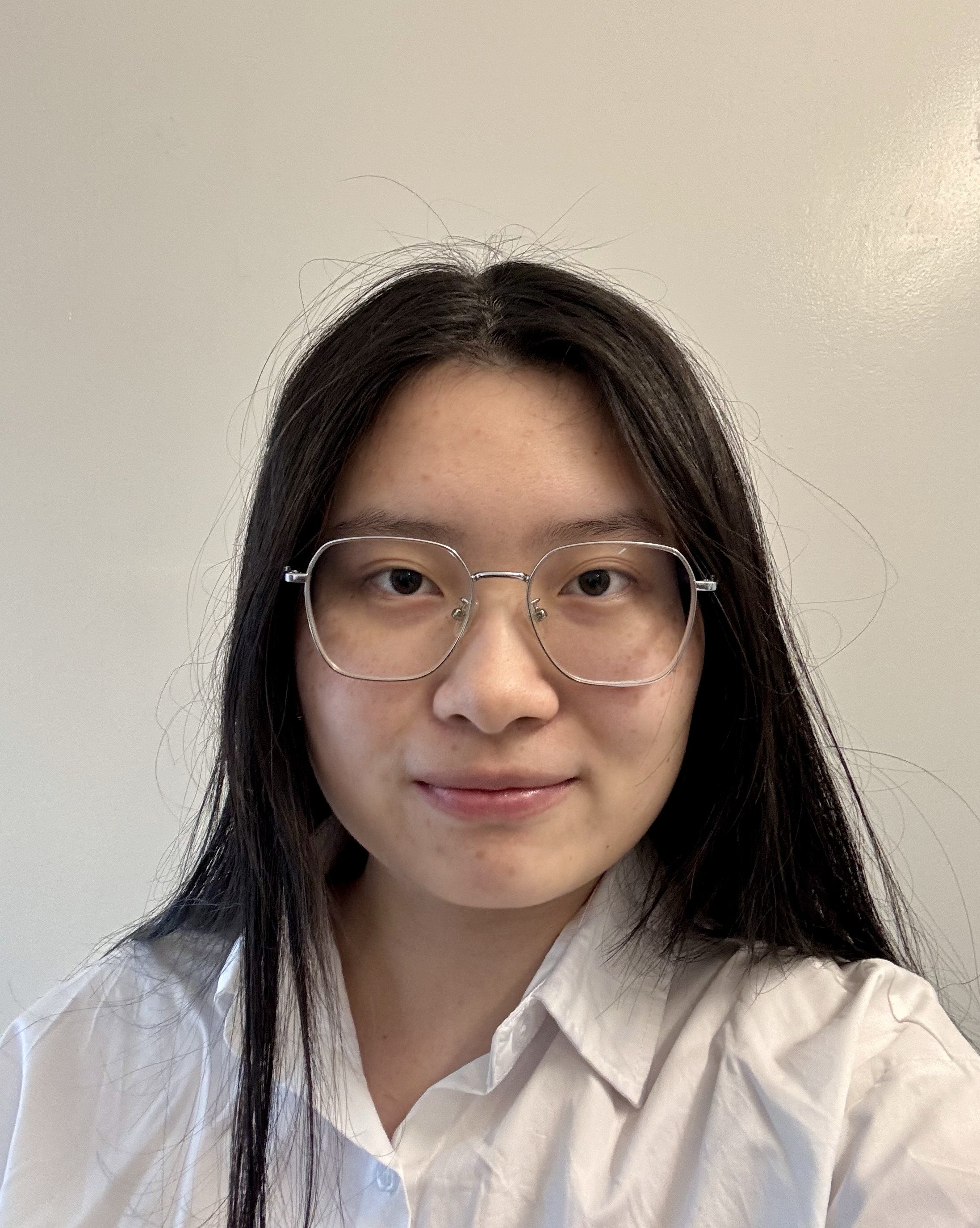 A young woman with long dark hair, glasses, and light skin wearing a white button-up shirt, looking at the camera against a plain wall background.
