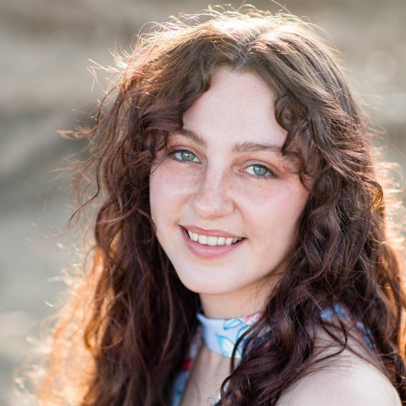 A young woman with long, wavy brown hair and blue eyes smiling at the camera, outdoors with sunlight in the background.