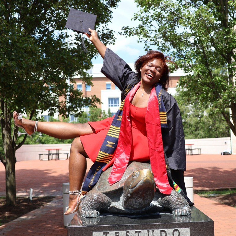A happy woman in a graduation gown and sash, sitting on a turtle statue outdoors, celebrating her graduation.