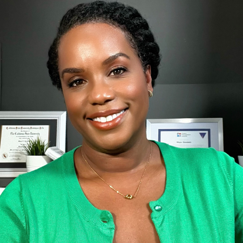 A smiling woman with short curly hair and brown skin, wearing a green cardigan and a gold necklace, standing in an office with framed certificates and a small potted plant in the background.