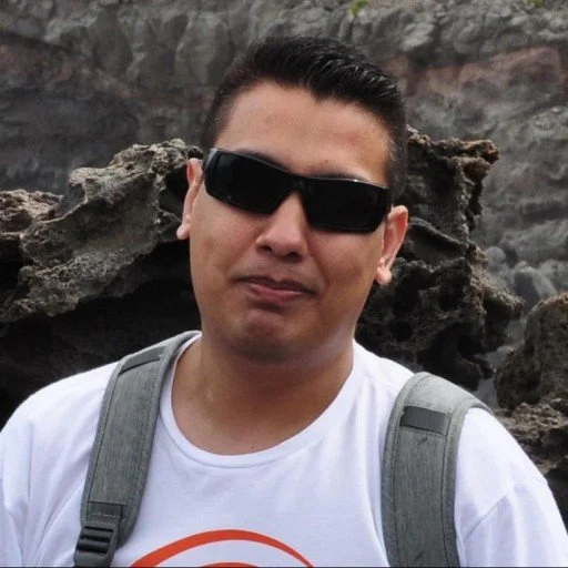 A man wearing sunglasses and a white t-shirt with a backpack standing outdoors with rocky terrain in the background.