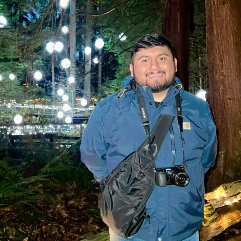 A man with a beard and short dark hair smiling outdoors at night in a forest, wearing a blue rain jacket with a camera hanging around his neck and a black camera bag hanging across his chest. Behind him, decorative hanging lights are illuminated among trees.