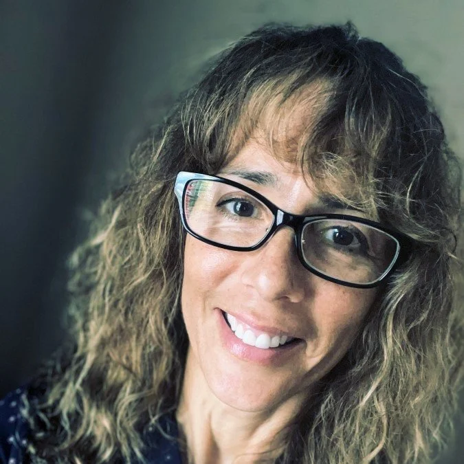 A woman with curly light brown hair and glasses smiling at the camera.