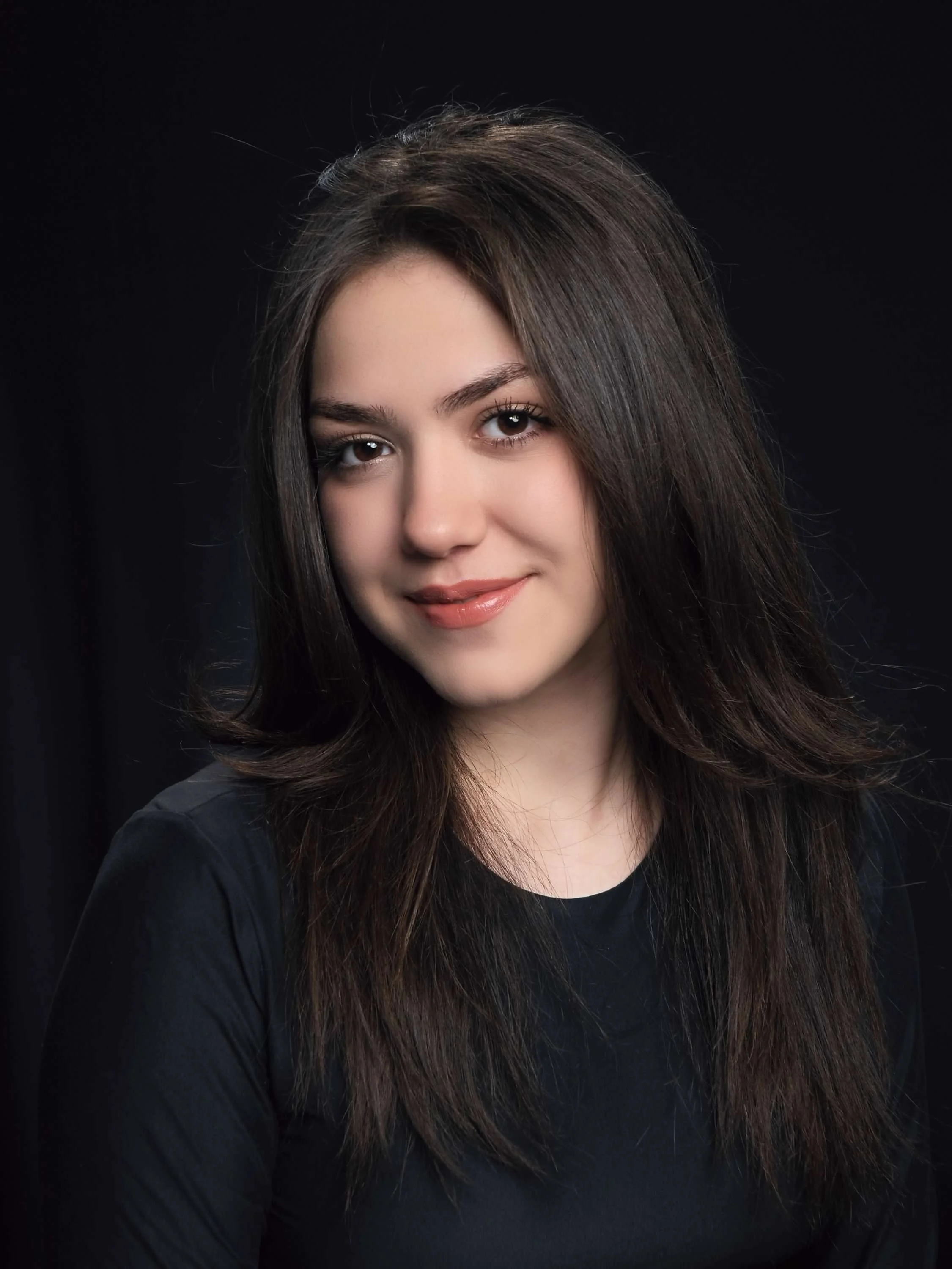 A man with a beard and short dark hair smiling outdoors at night in a forest, wearing a blue rain jacket with a camera hanging around his neck and a black camera bag hanging across his chest. Behind him, decorative hanging lights are illuminated among trees.