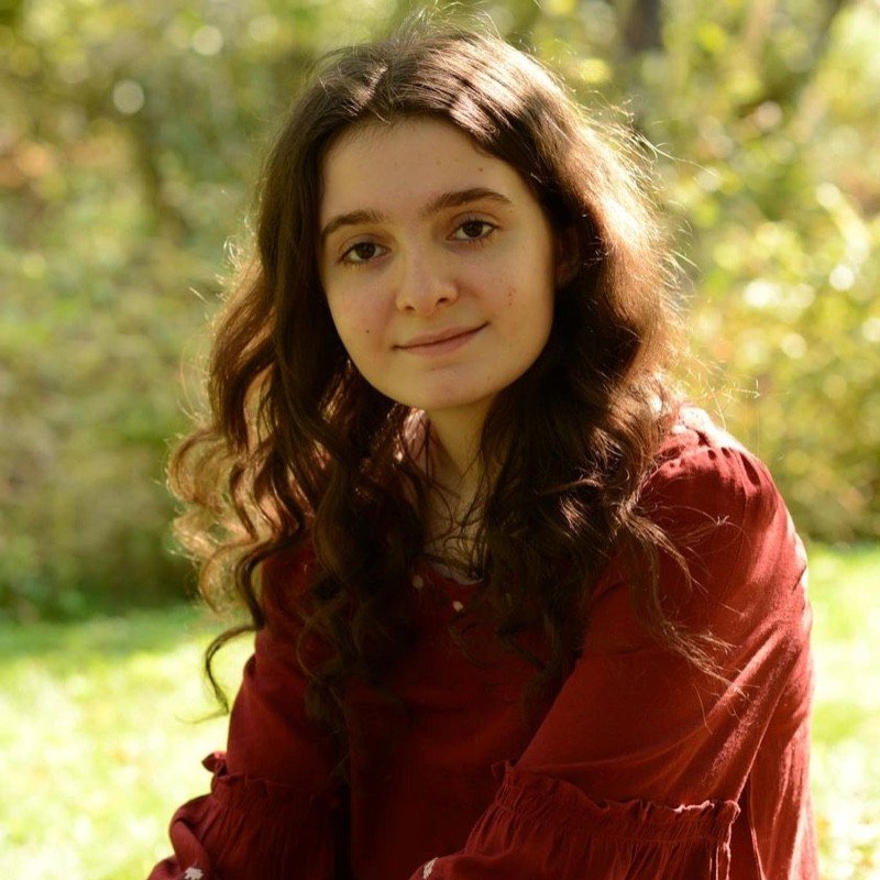 Young woman with long, curly brown hair, wearing a red dress, outdoors in a natural setting with green foliage in the background.