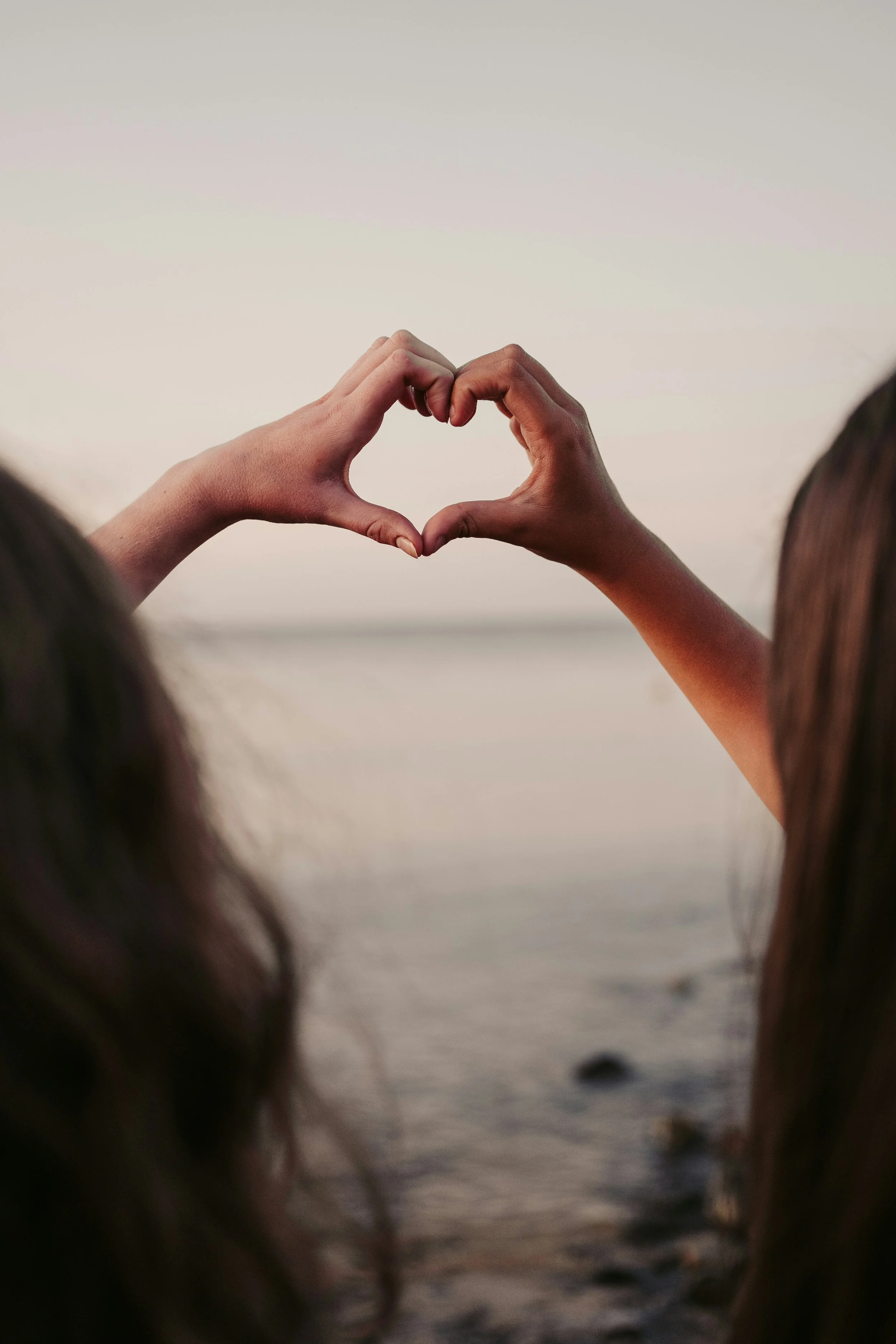 Two people forming a heart shape with their hands at the beach during sunset