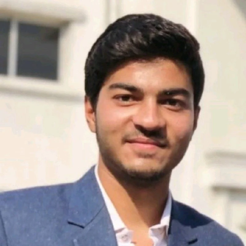 A young man with dark hair and a beard, wearing a blue blazer and white shirt, smiling in a well-lit indoor setting.