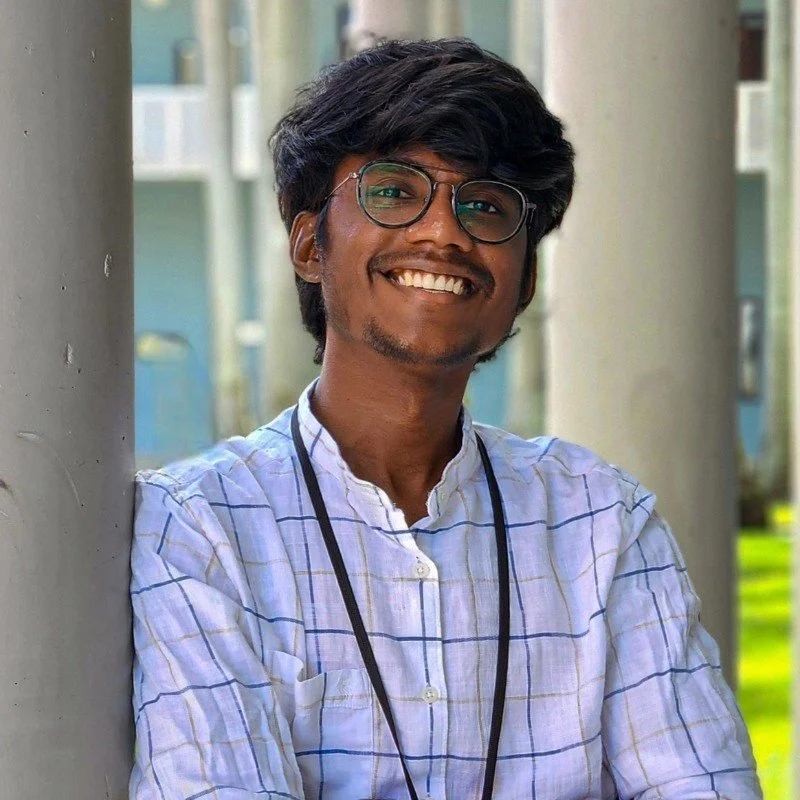A young man with dark hair and glasses, smiling and leaning against a pole outdoors with greenery in the background.