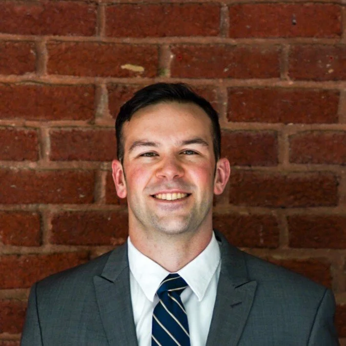 A man wearing a suit and tie, smiling, standing in front of a brick wall.