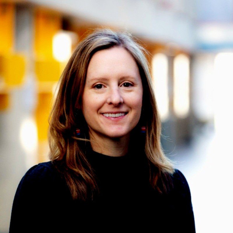 A smiling woman with shoulder-length brown hair, wearing a black top and earrings, standing in a well-lit indoor space with yellow and white accents in the background.