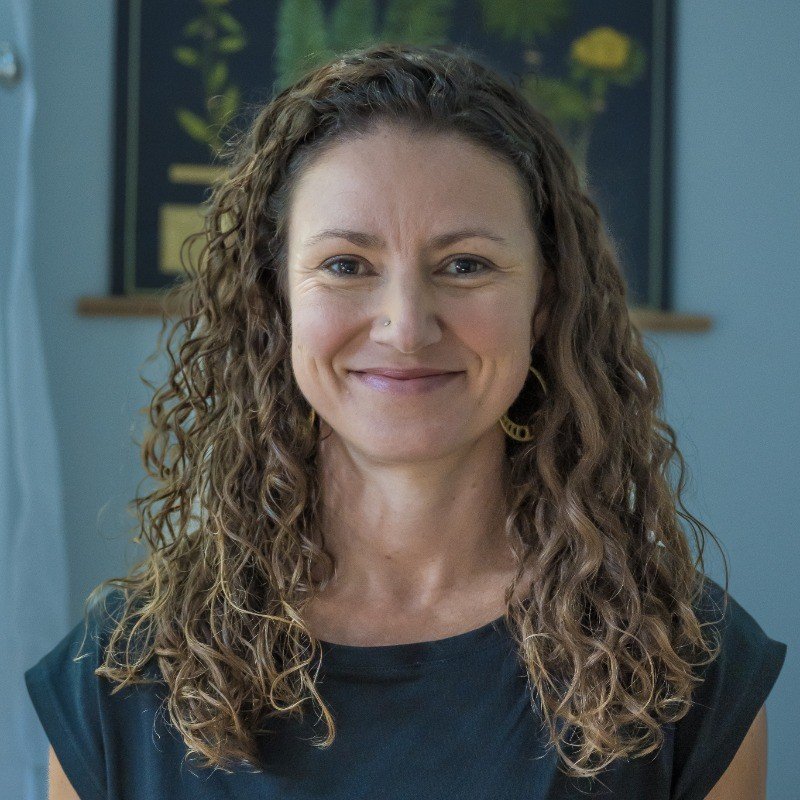 A woman with curly brown hair, smiling, wearing a black shirt and earrings, standing indoors with a framed picture and a plant in the background.