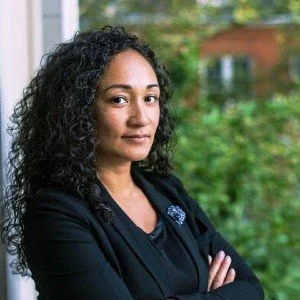 A woman with curly hair standing near a window with greenery outside.