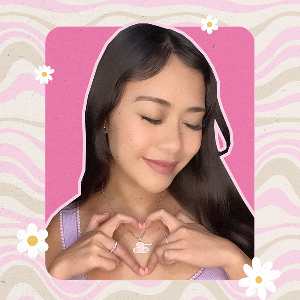 Young woman with long dark hair making a heart shape with her hands around a necklace with an Army Pink tank peace pendant, smiling softly with eyes closed, against a pink and beige wavy background with white daisies.