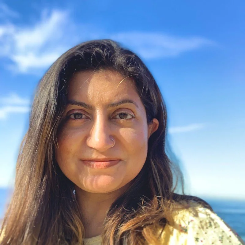 A woman with long brown hair, a nose piercing, and light makeup, smiling outdoors against a bright blue sky with some clouds, possibly near the coast.