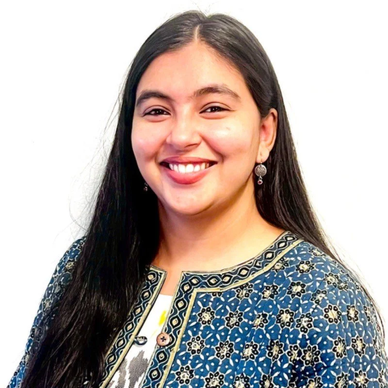 A young woman with long black hair smiling, wearing ornate earrings, and a blue patterned traditional jacket over a white top, standing against a white background.