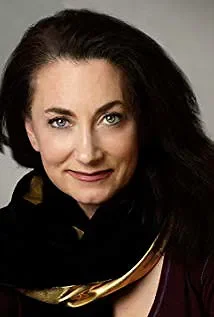 Close-up portrait of a woman with dark hair and blue eyes, wearing black with a hint of gold, smiling subtly against a gray background.
