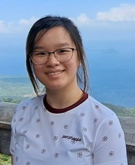 A young woman with long dark hair, wearing a black top, and smiling slightly, standing against a plain light-colored wall.