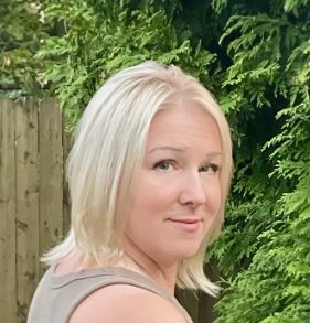 Portrait of a woman with blonde hair standing outdoors in front of green foliage and a wooden fence.