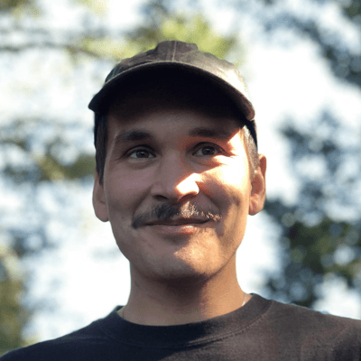 A man with a mustache wearing a black cap and black shirt, outdoors with blurred trees in the background.