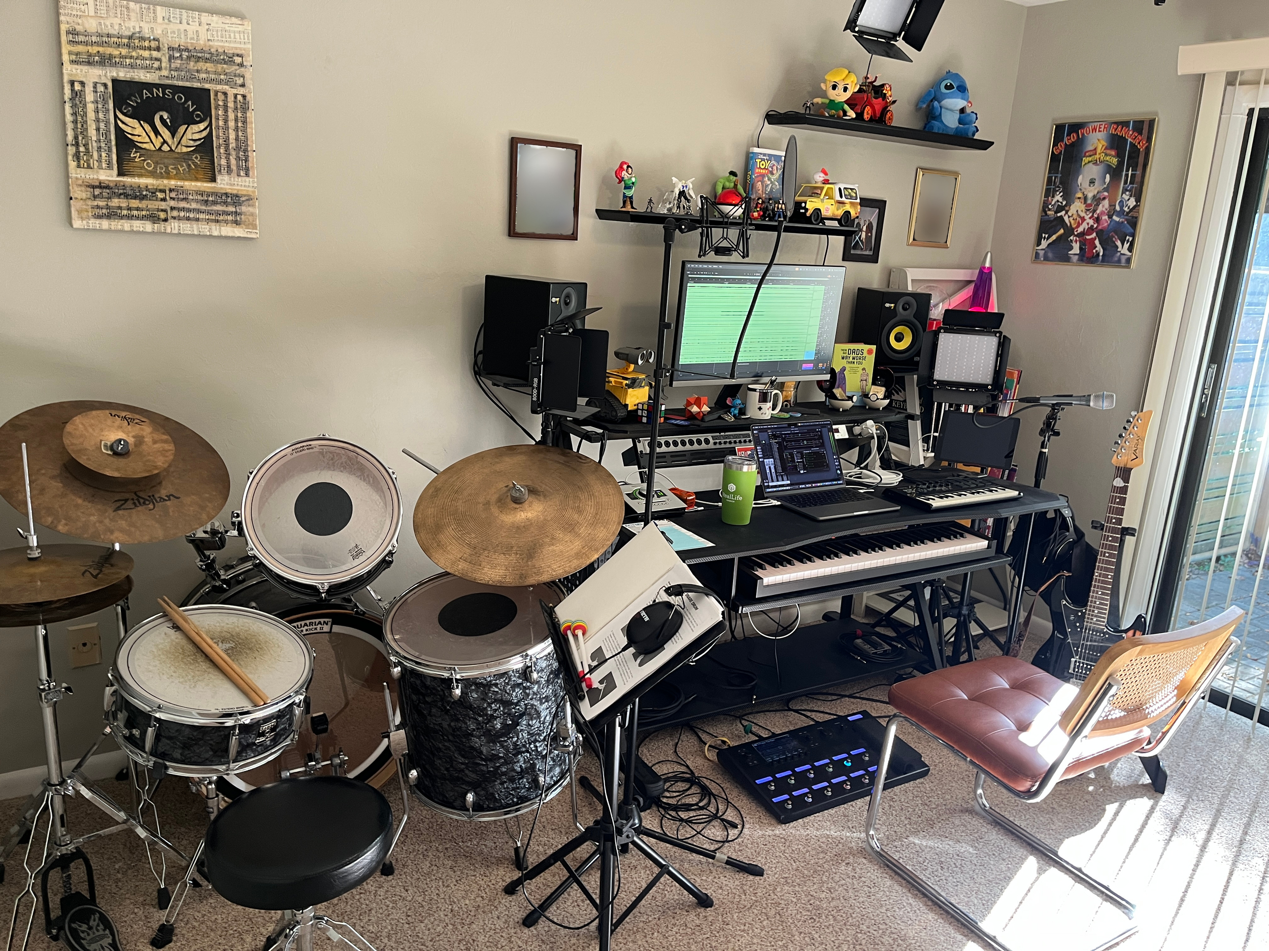 A music studio setup with a drum kit, a keyboard, a computer with audio software, speakers, and various toys and decorations on shelves and walls, including character figures from animation and a framed Power Rangers poster.