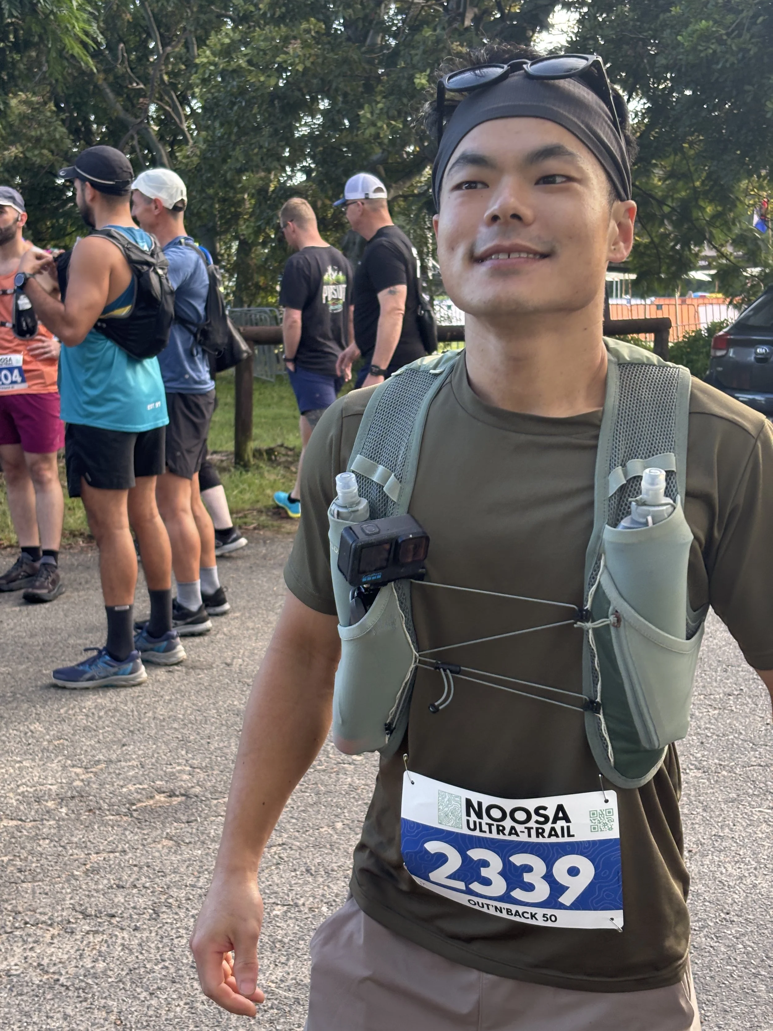 A young man participating in the NOOSA Ultra-Trail race, wearing a bib number 2339, a hydration vest with water bottles, and a headband with sunglasses on top, standing outdoors with other runners in the background.