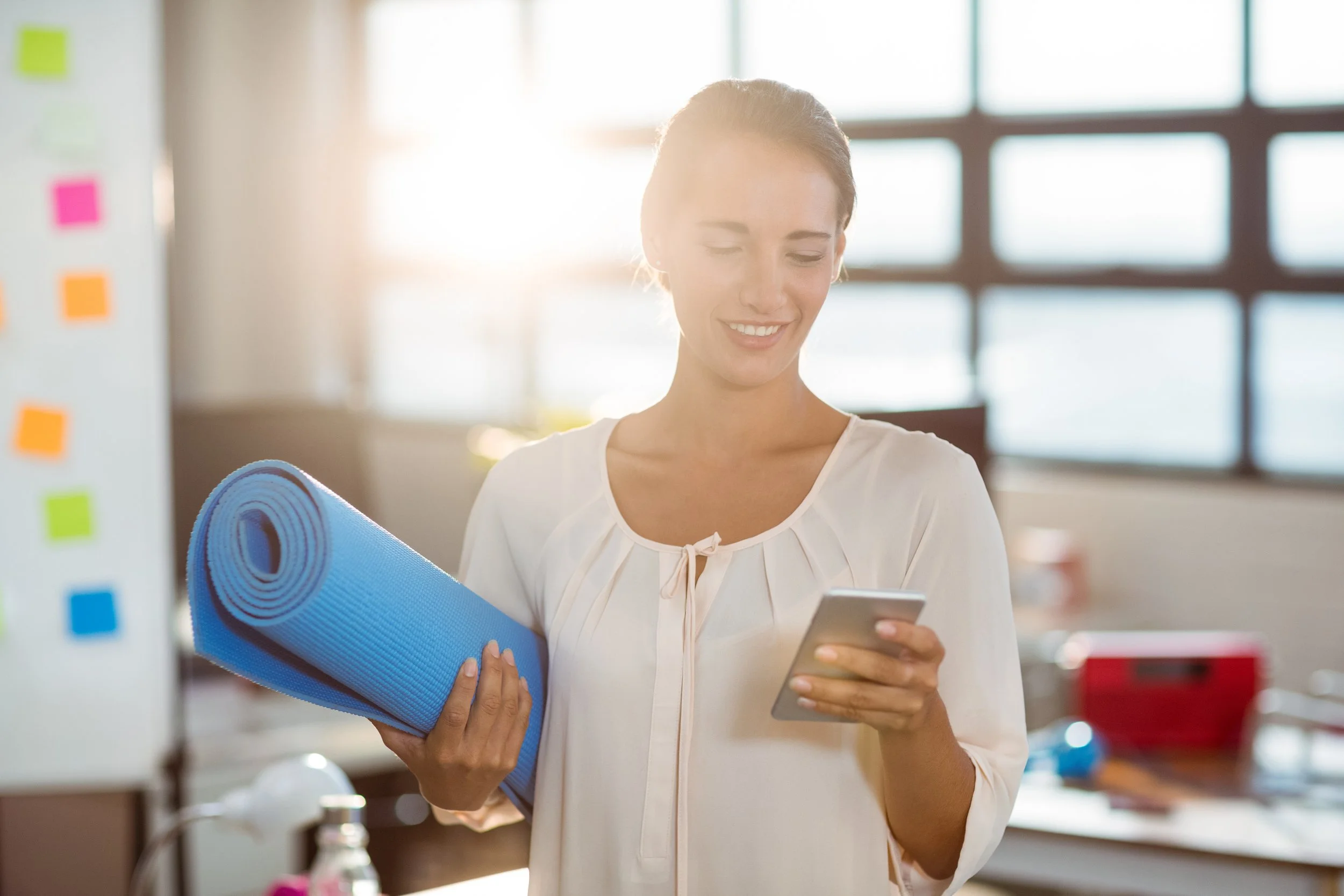 A woman smiling while looking at her phone, holding a blue yoga mat in a bright room with large windows. Booking now yoga classes. Book now.