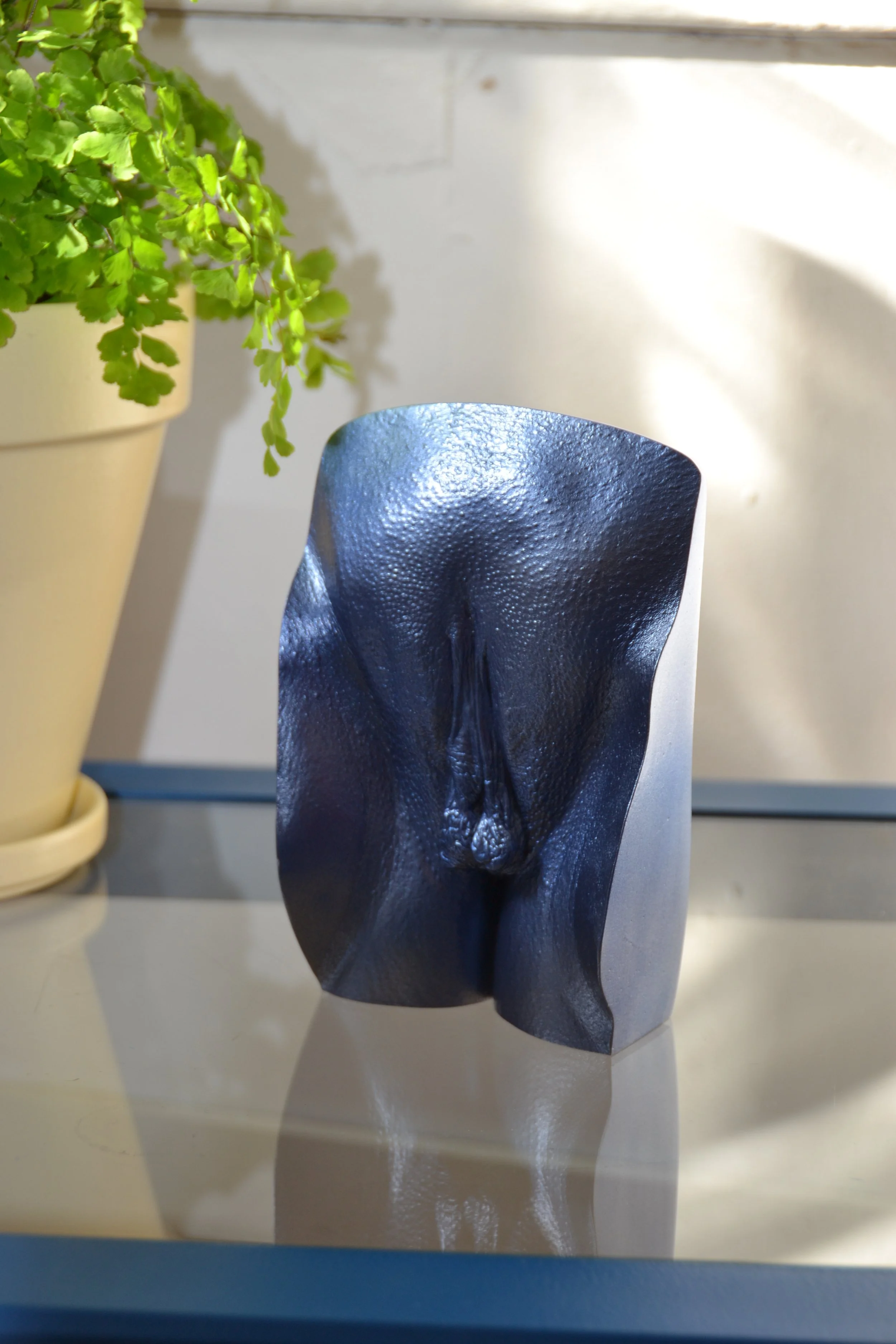 Metallic blauw vulva kunstwerk, handgemaakt van gips, geplaatst op een glazen tafel met een plant op de achtergrond, in atelier Venus in Vorm.