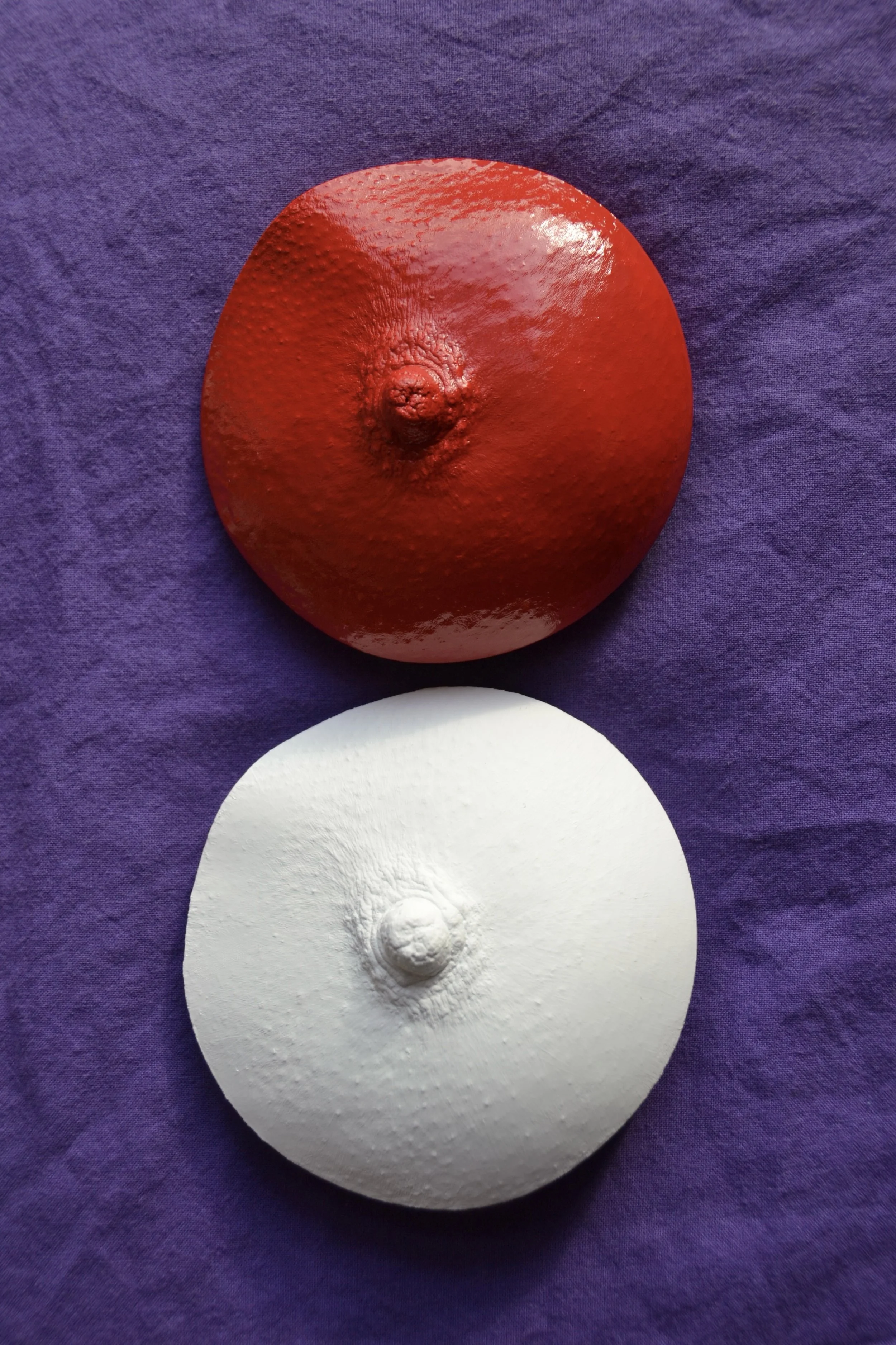 Twee ronde borstbeelden van gips in cirkelvorm, handgemaakt. De bovenste borst is rood en glanzend, de onderste wit en mat. Kunstwerk van Venus in Vorm, gefotografeerd op donkerpaarse stof.
