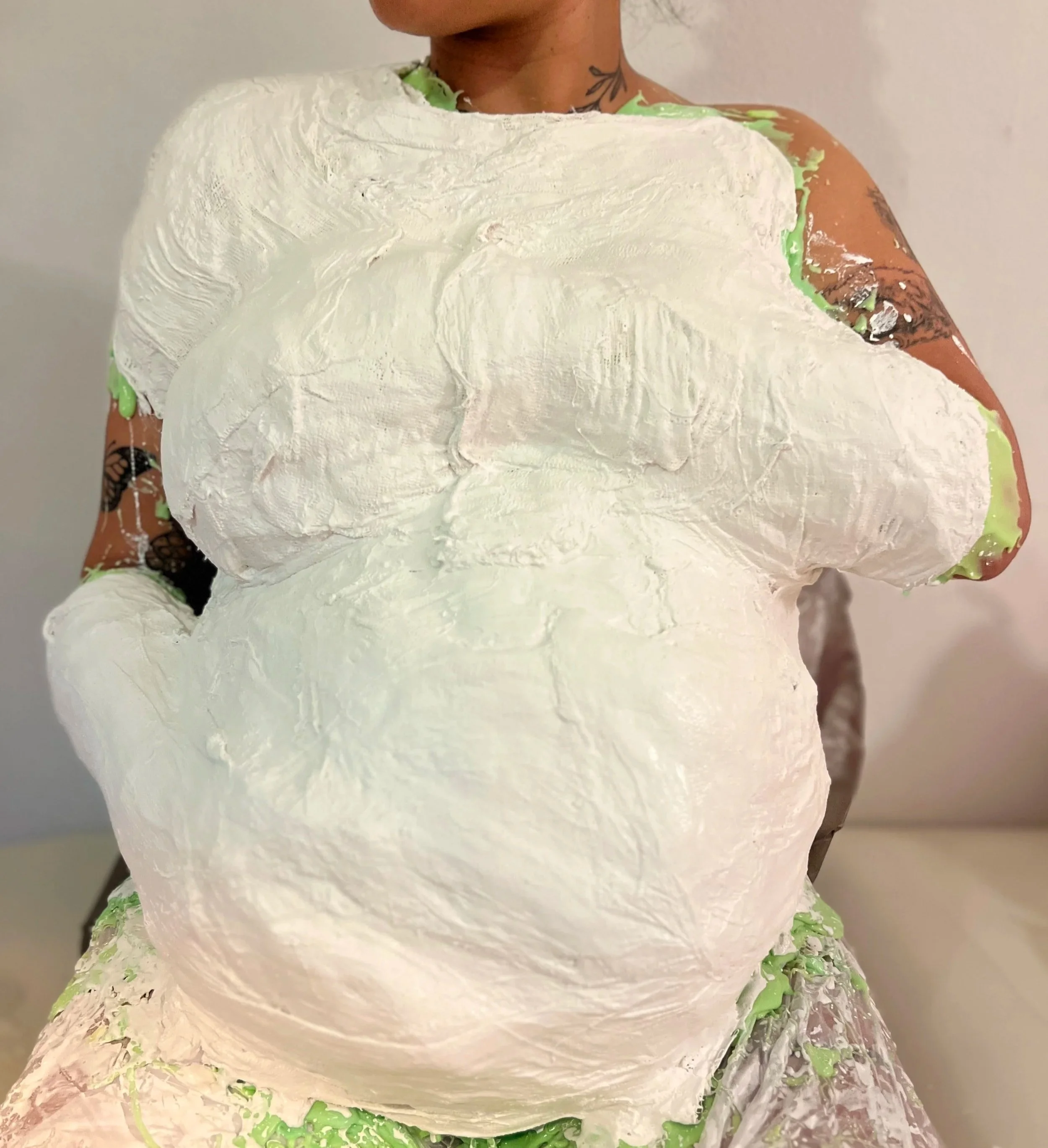 Vrouw zittend in atelier van Venus in Vorm tijdens body casting, bedekt met groene siliconen en een witte steunkap van gips. Tattoos zichtbaar op hals en armen.