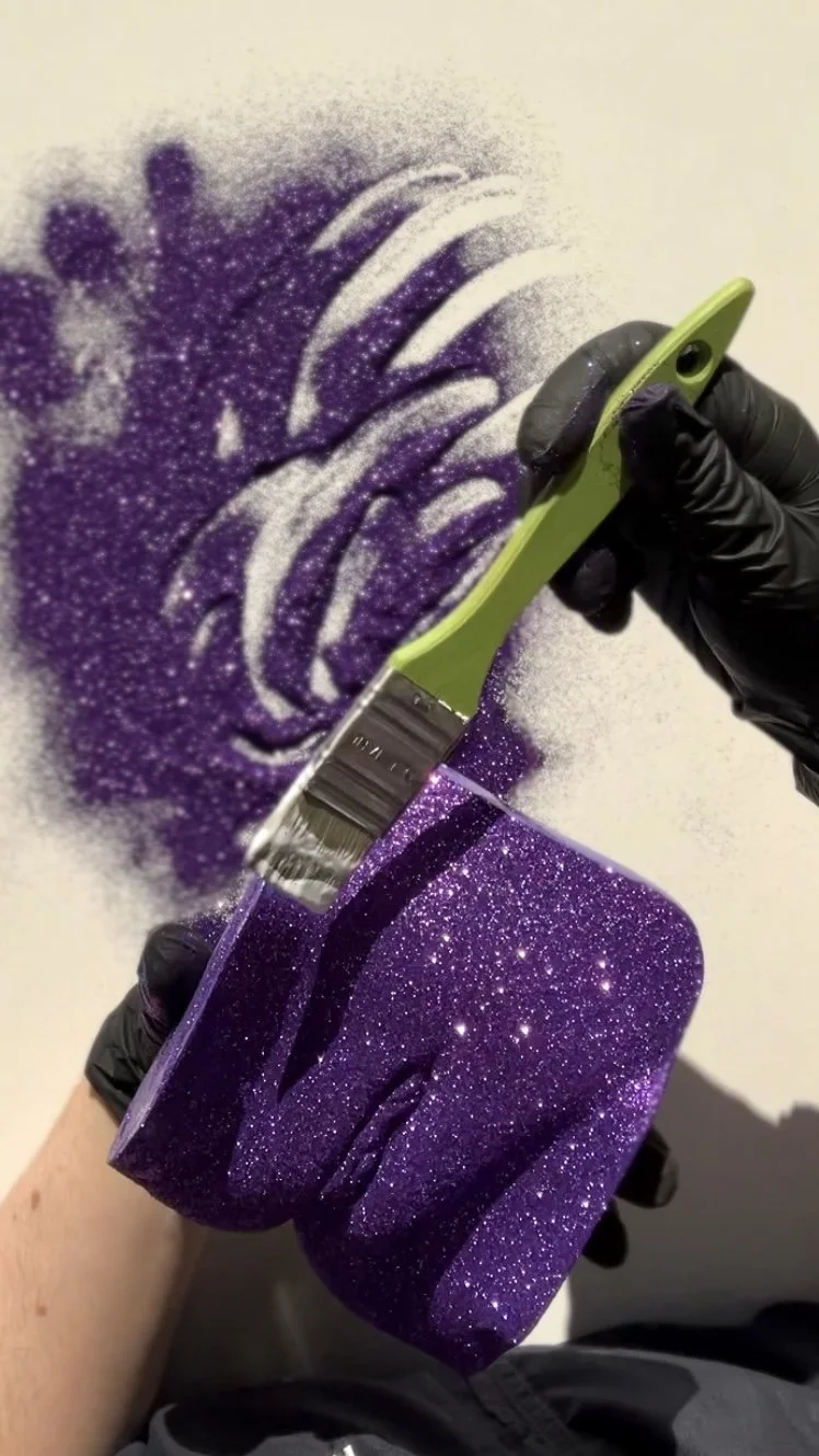 Werk in uitvoering: handgemaakt vulva-beeld van gips met paarse glitters, glitters aangebracht door de kunstenaar met handschoentjes aan, kunstwerk van Venus in Vorm, gefotografeerd op witte tafel in het atelier in zonlicht.