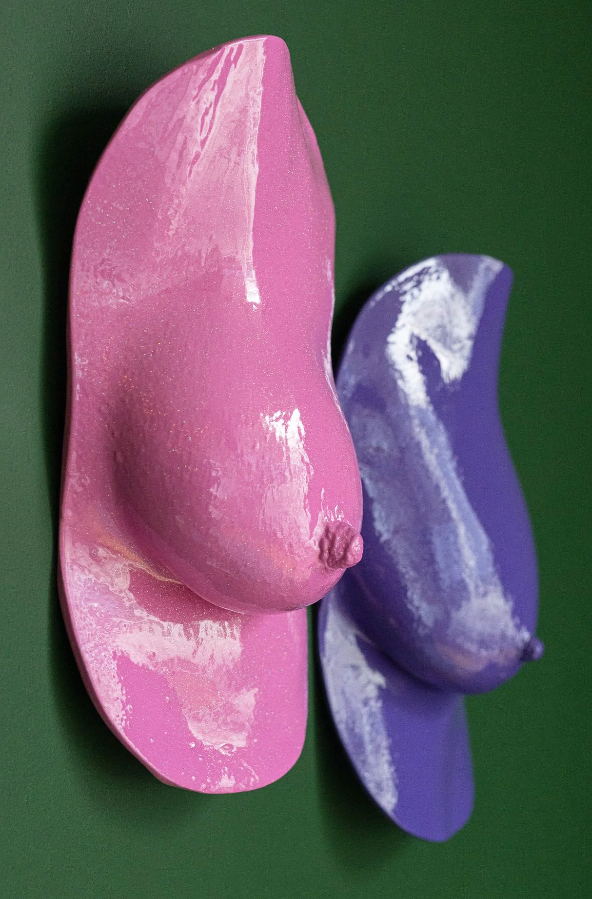 Zijaangezicht van twee borstsculpturen in roze en paars tegen een groene achtergrond, handgevormd naar echte lichamen.